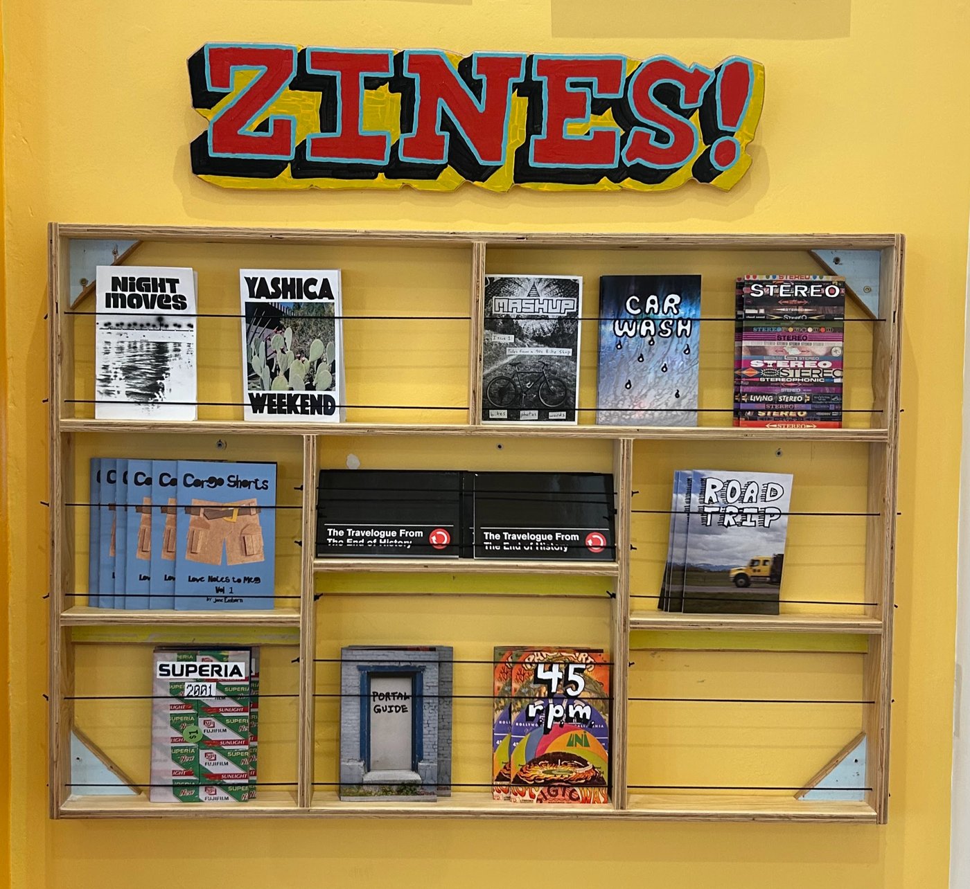 The ZINES! display at the Zootown Arts Community Center: a light wood wall shelf with cords holding Mashup Publishing zines and work by other local zine makers